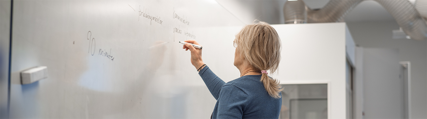 En lärare med ljust hår och hästsvans står och skriver på en whiteboard.